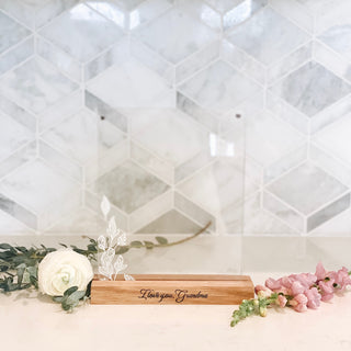 "I love you, Grandma" written in cursive engraved in a wood base picture frame holding up two pieces of acrylic with leaves on the left side and the frame is surrounded by two flowers on a white kitchen counter