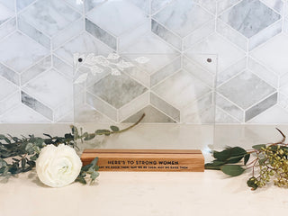 "Here's to strong women may we know them, may we be them, may we raise them" engraved in black ink on a wooden base holding two pieces of acrylic with leaves carved into the top left in white. the frame itself is centered between two flowers on a white kitchen counter with a marble backsplash