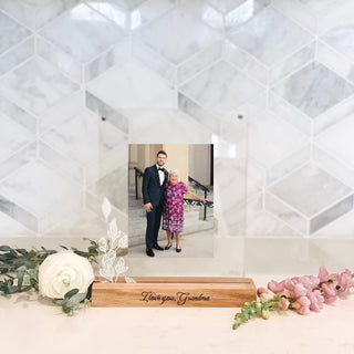 "I love you, Grandma" written in cursive engraved in a wood base picture frame holding up two pieces of acrylic with leaves on the left side holding a photo of an older grandson in a suit and his grandma in a purple dress. the frame comes in various sayings