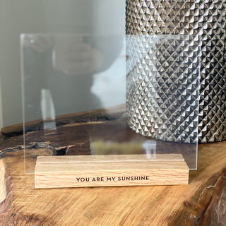 "you are my sunshine" carved into a wood base frame in blank ink with two pieces of acrylic to hold a picture. frame is on a natural wood table with a metallic studded vase behind it